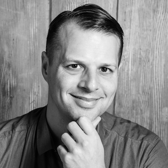 Black and white portrait of a man with slicked-back hair, smiling, with his hand resting on his chin, against a wooden background.