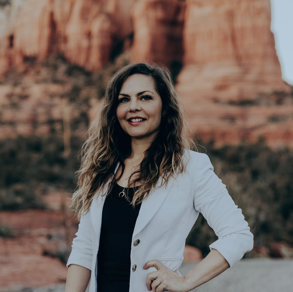 A woman with long, wavy brown hair wearing a black top and a white blazer smiling outdoors with red rock formations in the background.