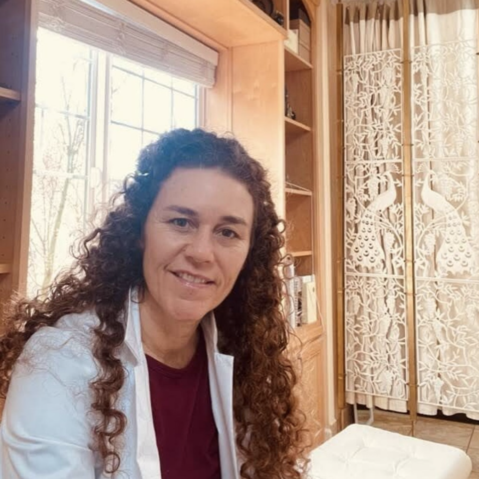 A woman with curly hair sitting in a room with wooden shelves, a window, and decorative peacock and nature-themed carved screens.