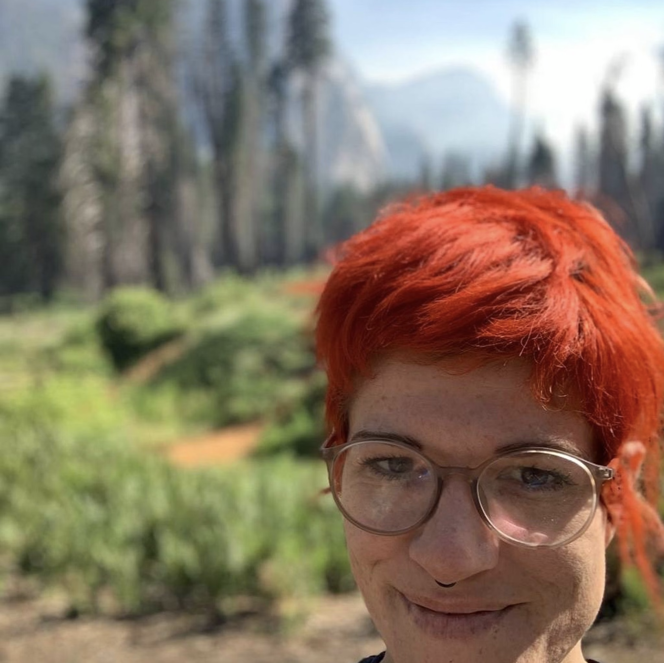 Close-up of a person with bright red hair, wearing glasses, smiling outdoors with a forest background.