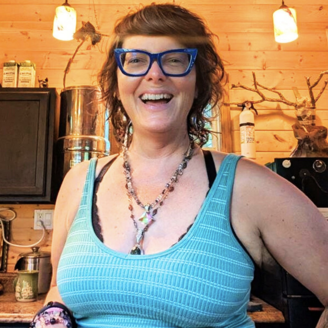 A smiling woman with curly red hair and blue glasses wearing a turquoise tank top and layered necklaces, standing in a warmly lit wooden interior.
