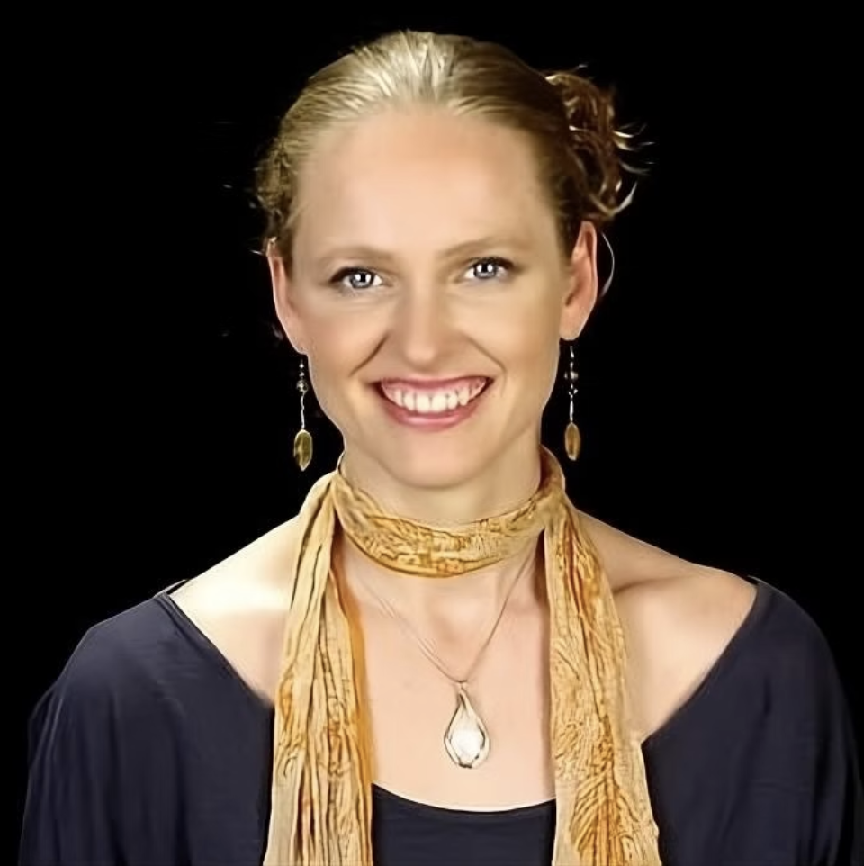 Portrait of a smiling woman with blonde hair pulled back, wearing earrings, a gold scarf on her neck, and a necklace with a teardrop pendant, against a black background.
