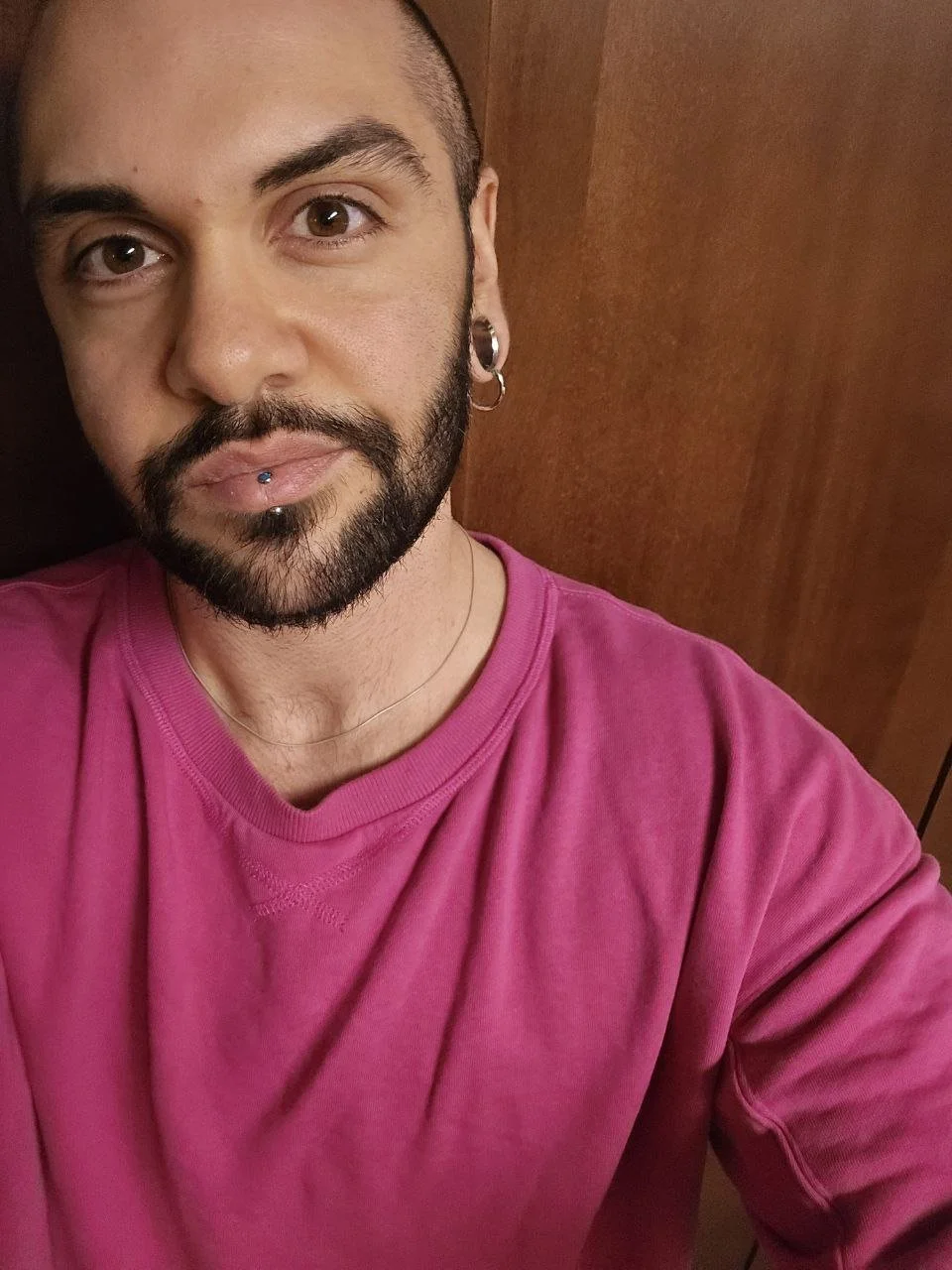 A man with a beard and piercings wearing a pink shirt, taking a selfie indoors against a wooden background.