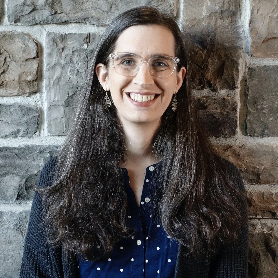 A woman with long, wavy dark hair and glasses smiling in front of a stone wall, wearing silver earrings and a navy blue polka-dot shirt.
