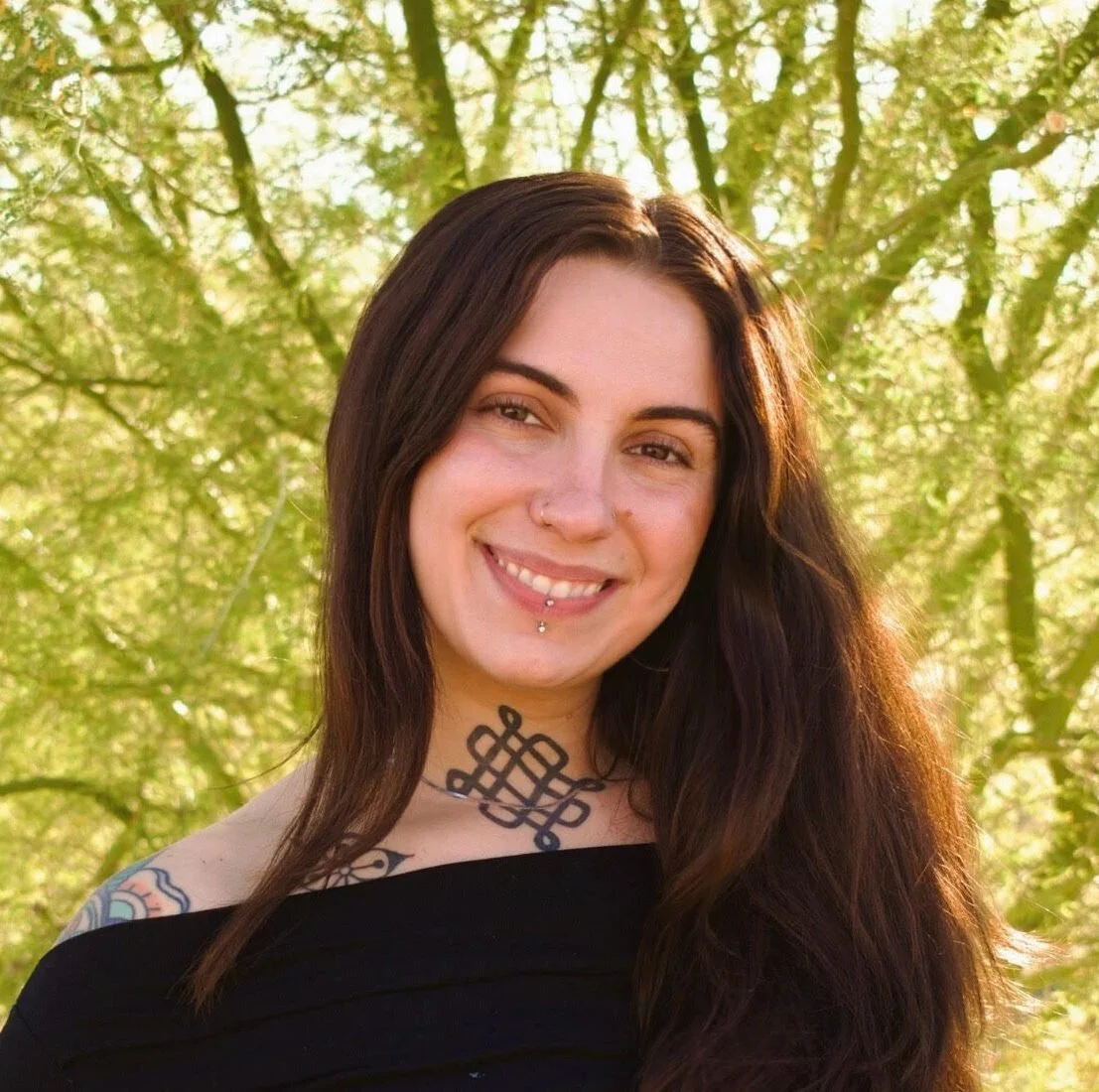 A smiling woman with long dark hair and tattoos, standing outdoors with green trees in the background.