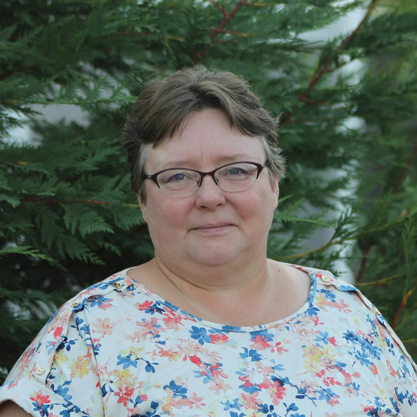 A woman with short brown hair and glasses standing in front of green foliage, wearing a floral blouse.