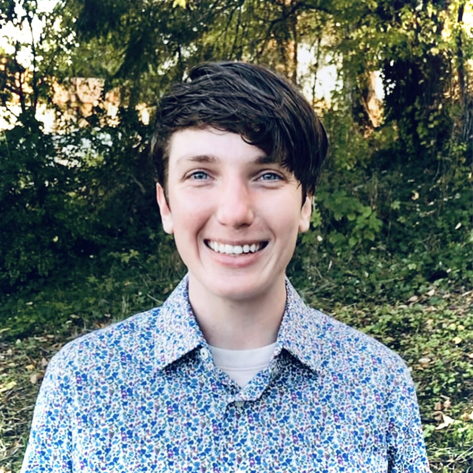 A young man with dark hair and blue eyes smiling outdoors with trees and greenery in the background.