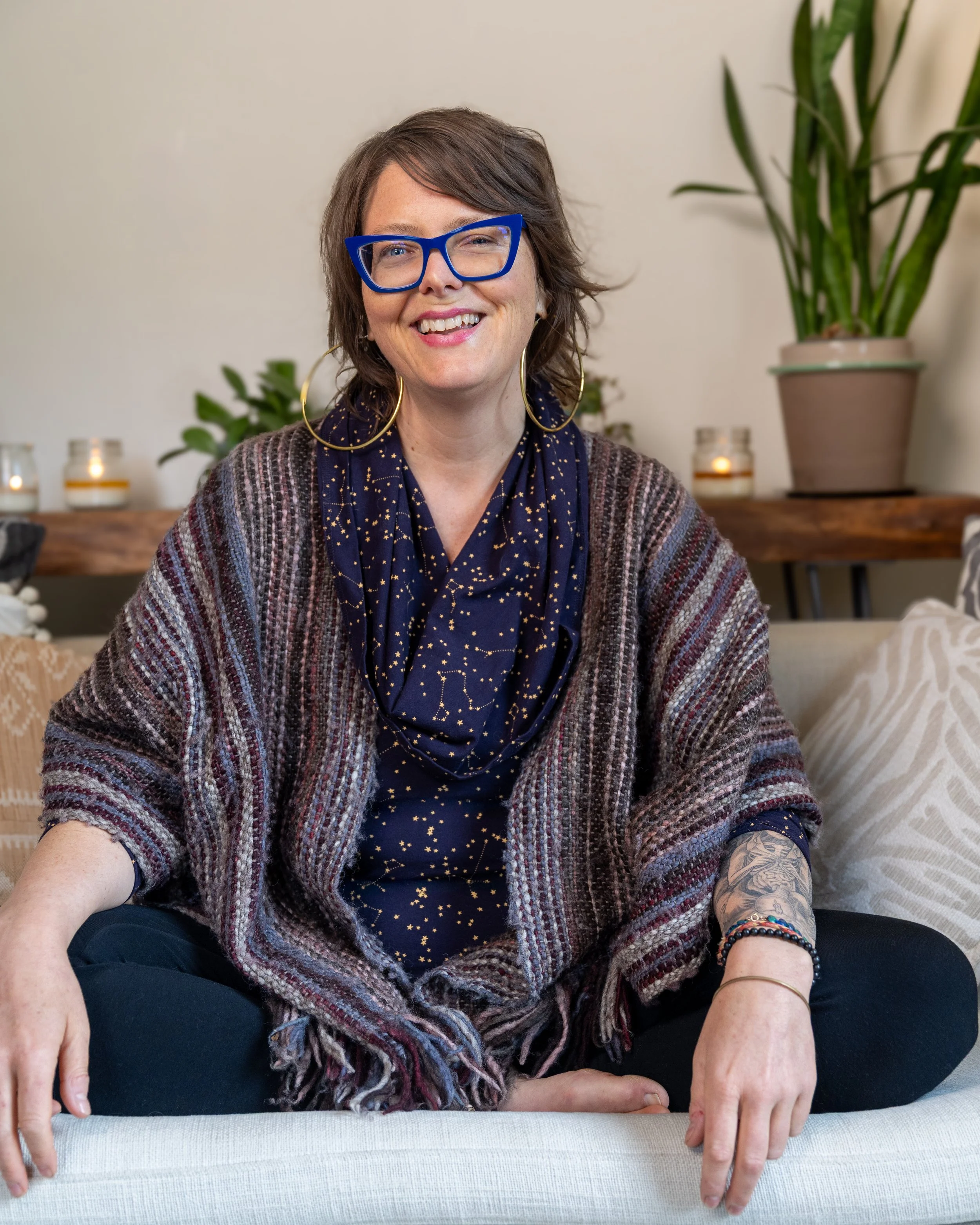 A person with short brown hair and blue glasses smiling, wearing large gold hoop earrings and a dark scarf with a star pattern, standing in front of a neutral background with greenery.