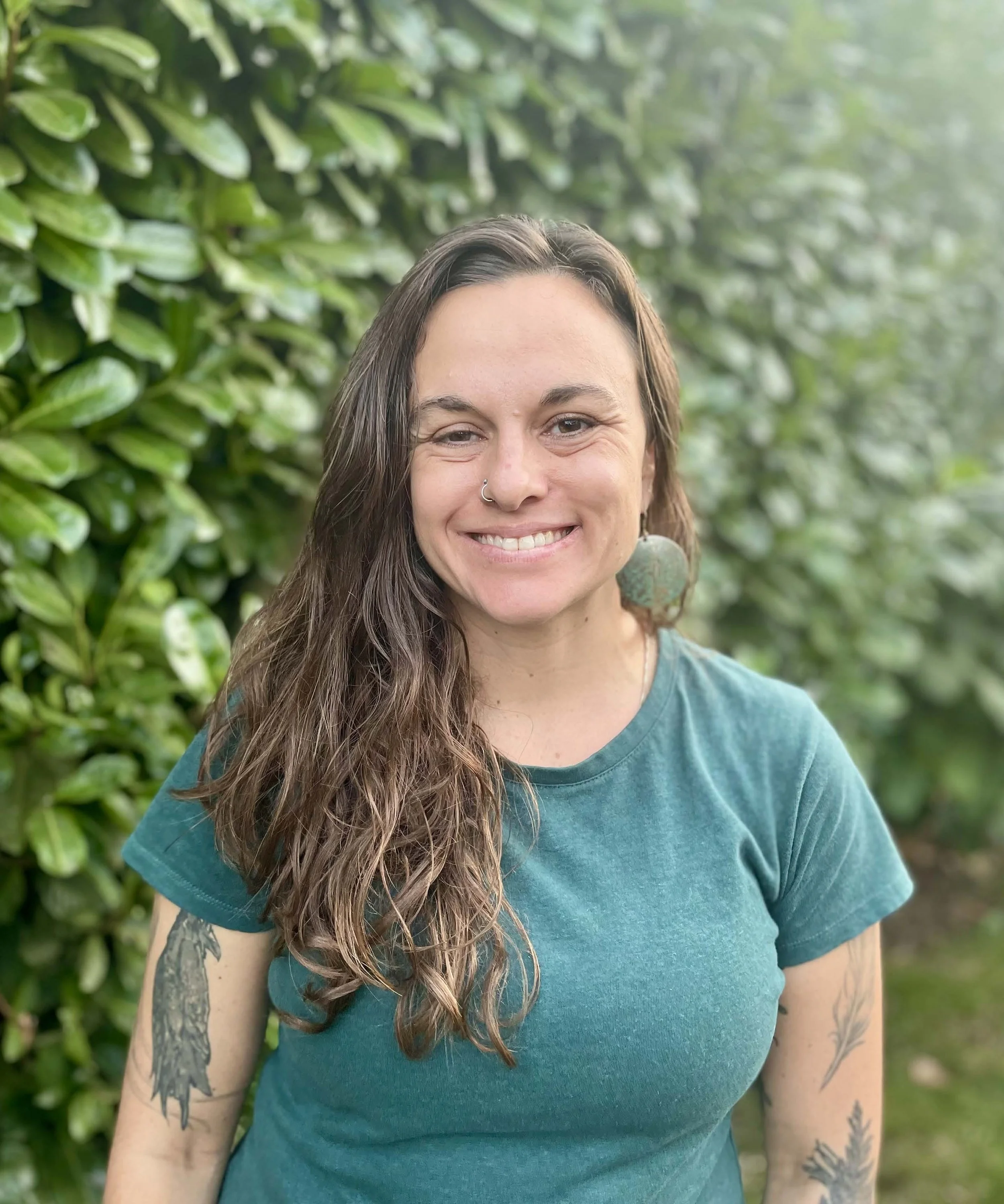 A smiling woman with long wavy brown hair, wearing a teal T-shirt and large earrings, standing outdoors in front of green foliage.