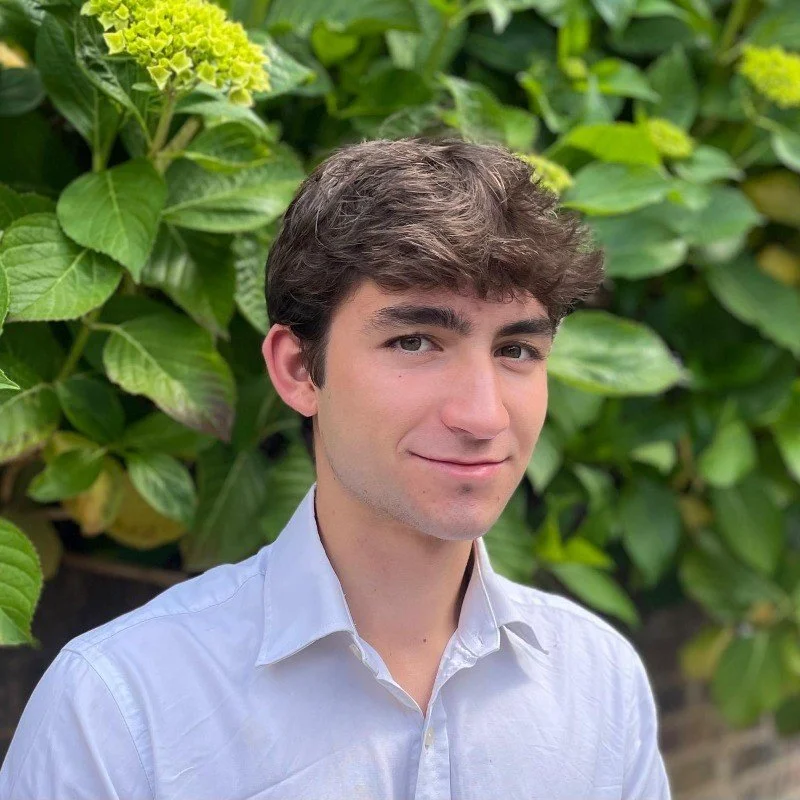 Young man with brown hair and light skin, wearing a white collared shirt, outdoors with green foliage in the background.