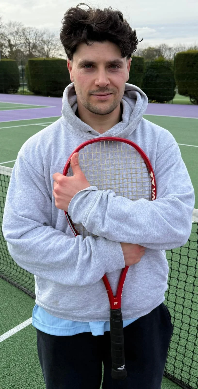 A man standing on a tennis court holding a tennis racket, wearing a gray hoodie and black pants, with trees and a net behind him.