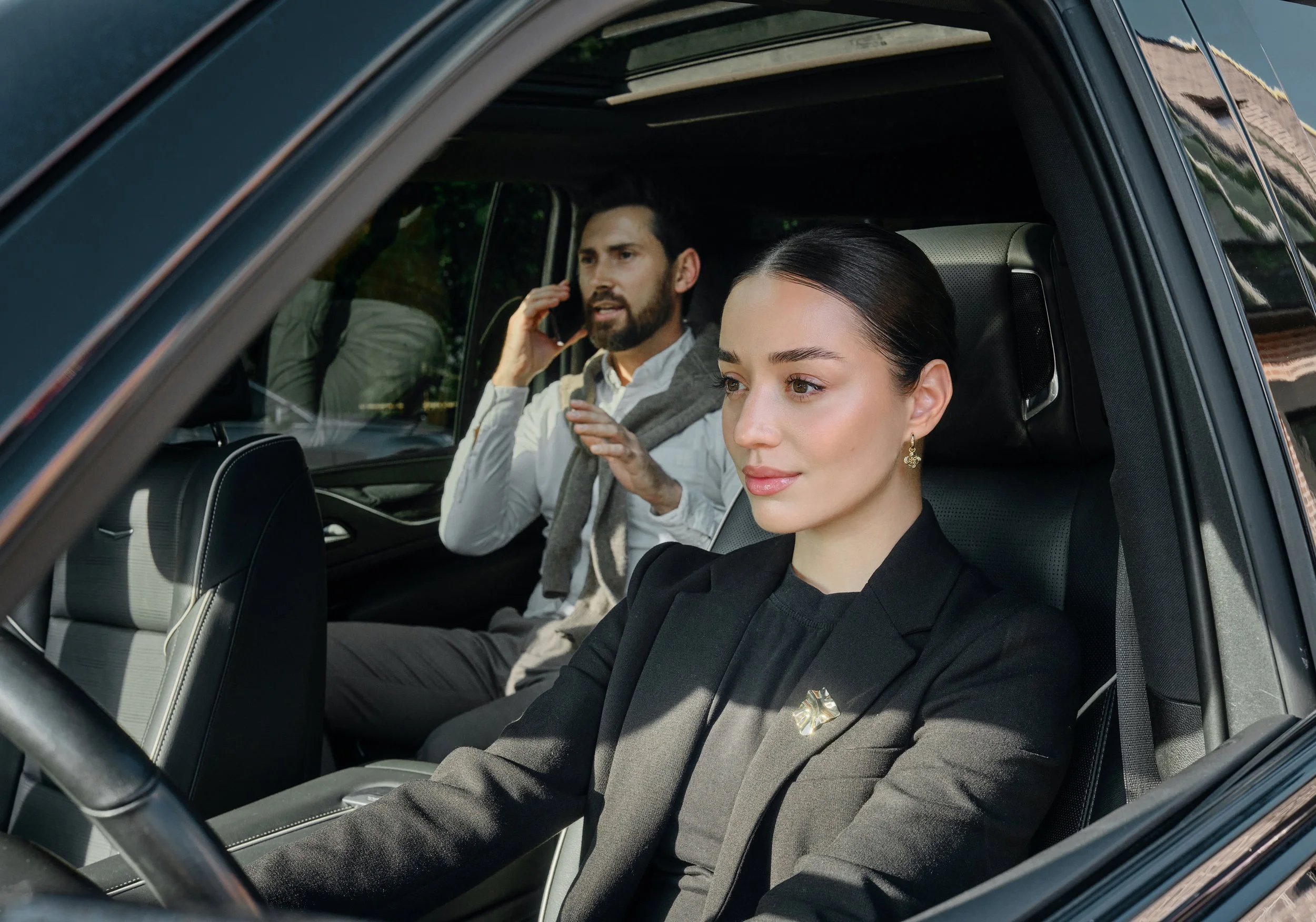 A woman in a black blazer sitting in the driver's seat of a car, with a man in a white shirt and scarf in the passenger seat talking on a phone.
