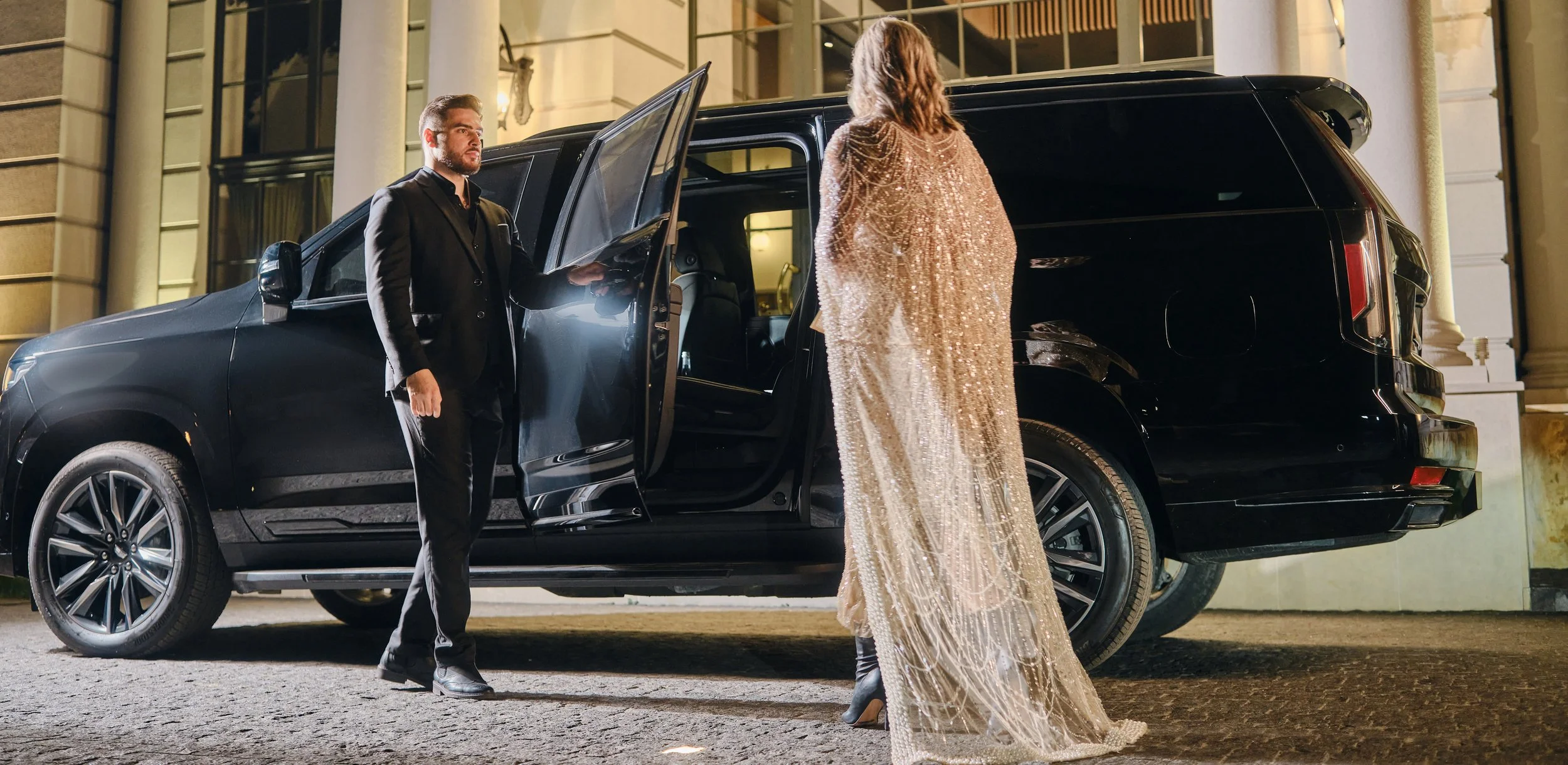 A woman in a sparkling, gold evening gown is approaching a black luxury SUV parked outside a building, as a man in a black suit opens the door for her at night.