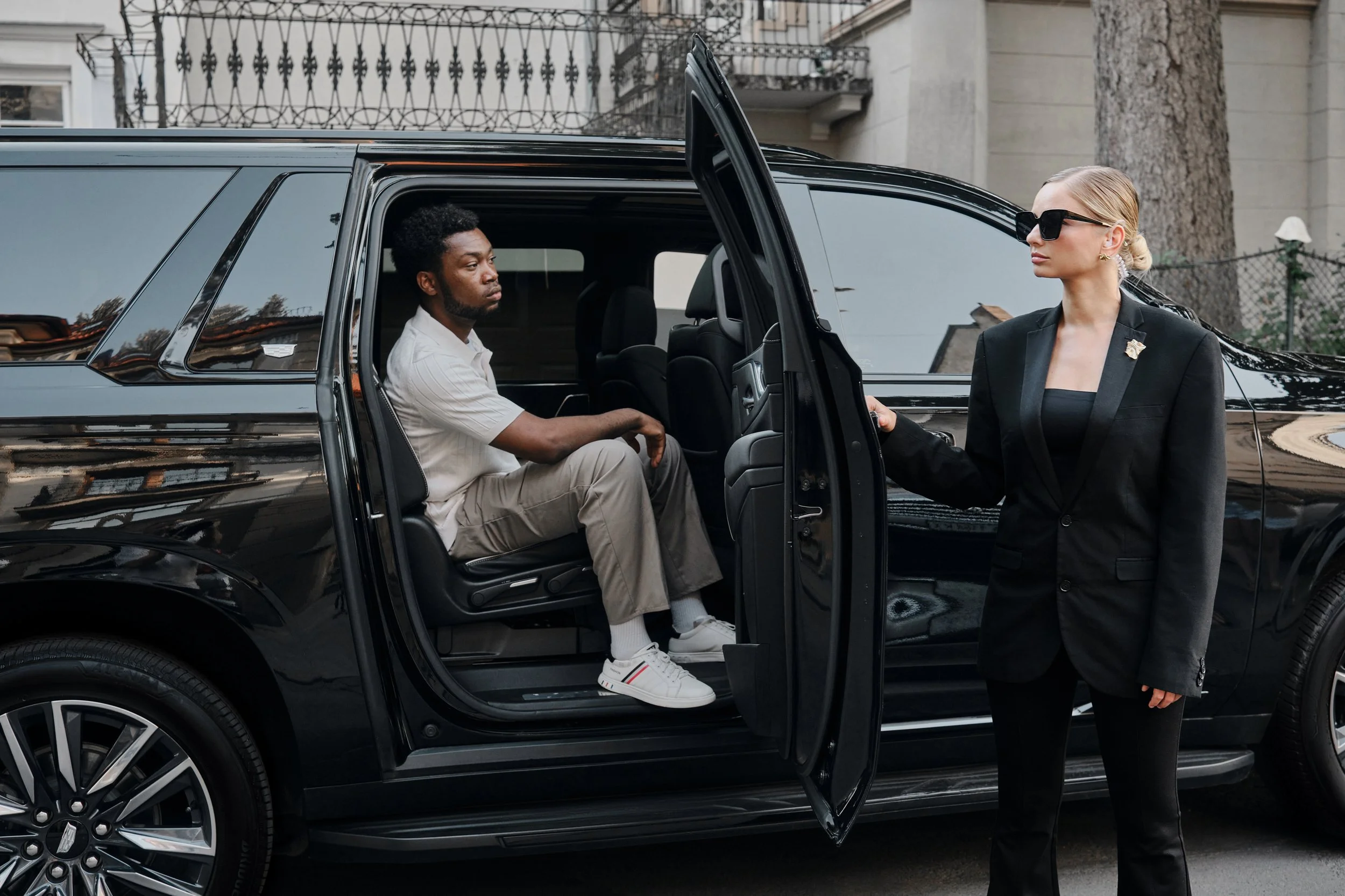 A woman in a black suit and sunglasses standing outside of a black limousine, holding the door open for a man sitting inside, wearing a beige outfit.