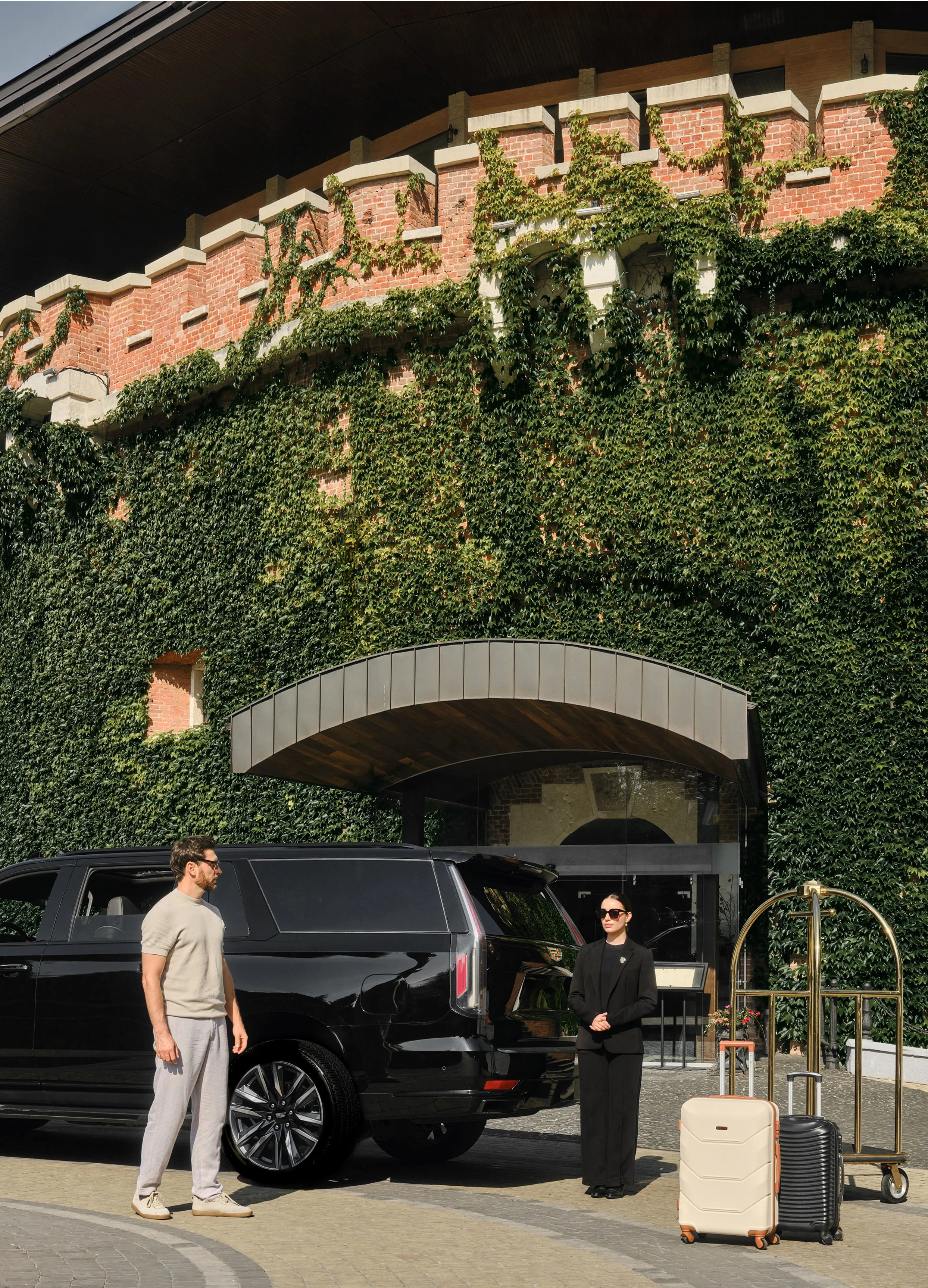 A man and a woman stand outside a hotel with dark suitcases near a luxury black vehicle. The hotel has a steep brick wall covered in green ivy and a glass entrance with a curved awning.