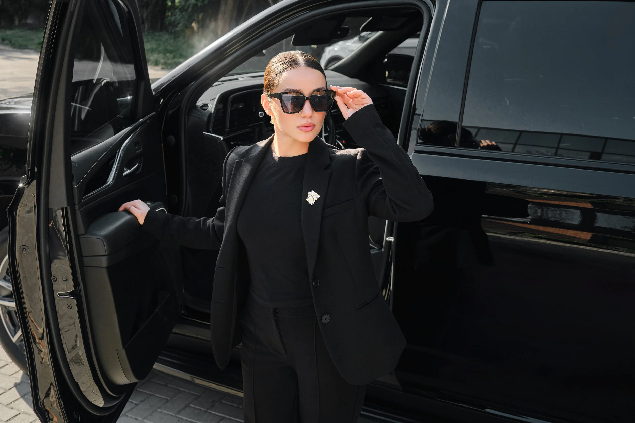 A woman dressed in black, wearing sunglasses, standing beside a black luxury SUV with the door open, adjusting her glasses.