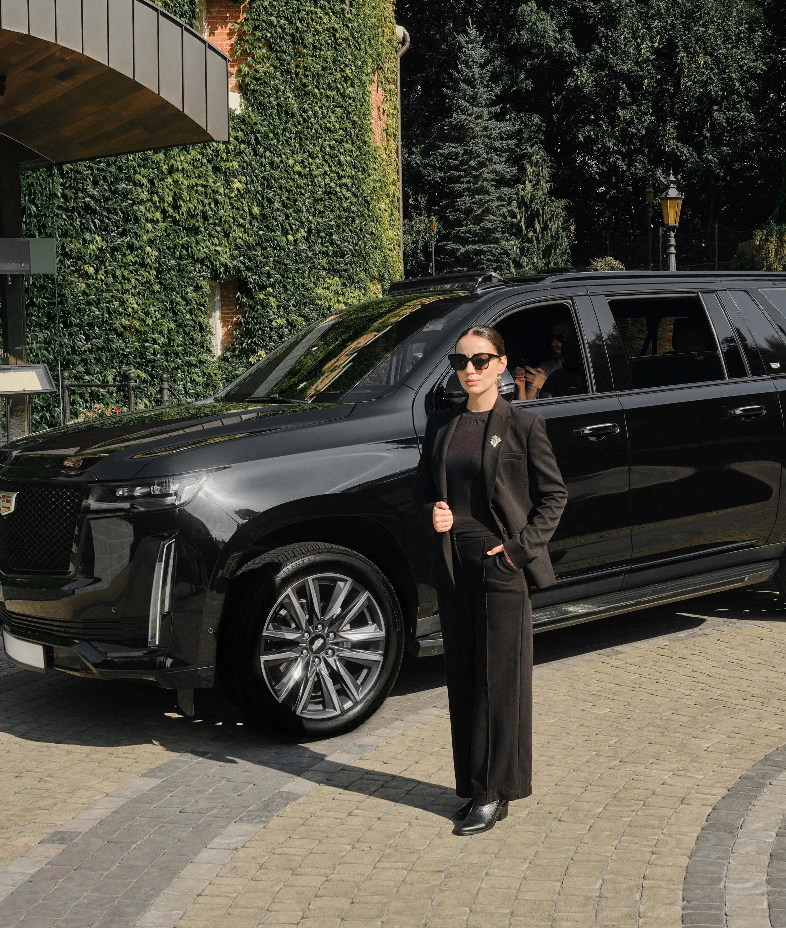 A woman in sunglasses and a black blazer stands next to a black luxury SUV, with a person visible inside the vehicle using a mobile phone, in a setting with green ivy-covered walls and trees.