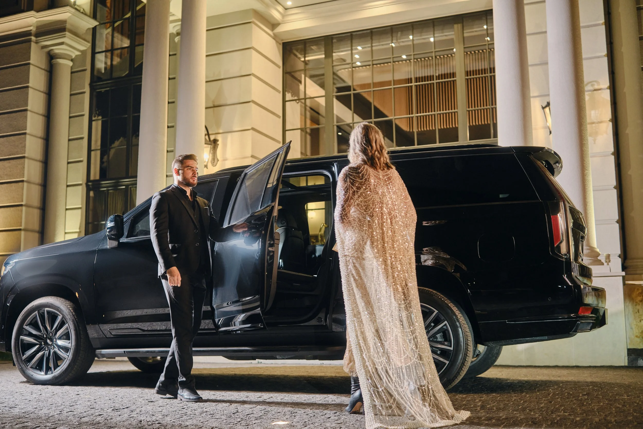 A woman in a gold sequined dress standing outside a black SUV at night, talking to a man in a black suit as he holds the car door open, in front of a large, elegant building with columns and tall windows.