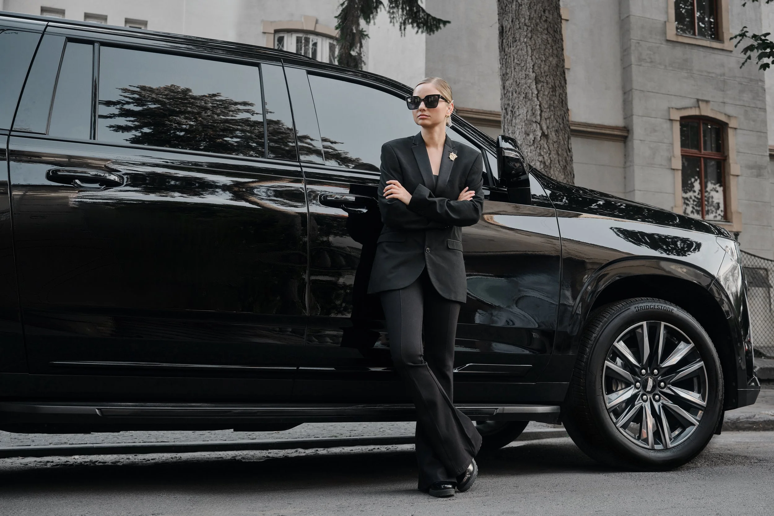 A woman wearing sunglasses and a black blazer standing next to a black SUV on a city street.