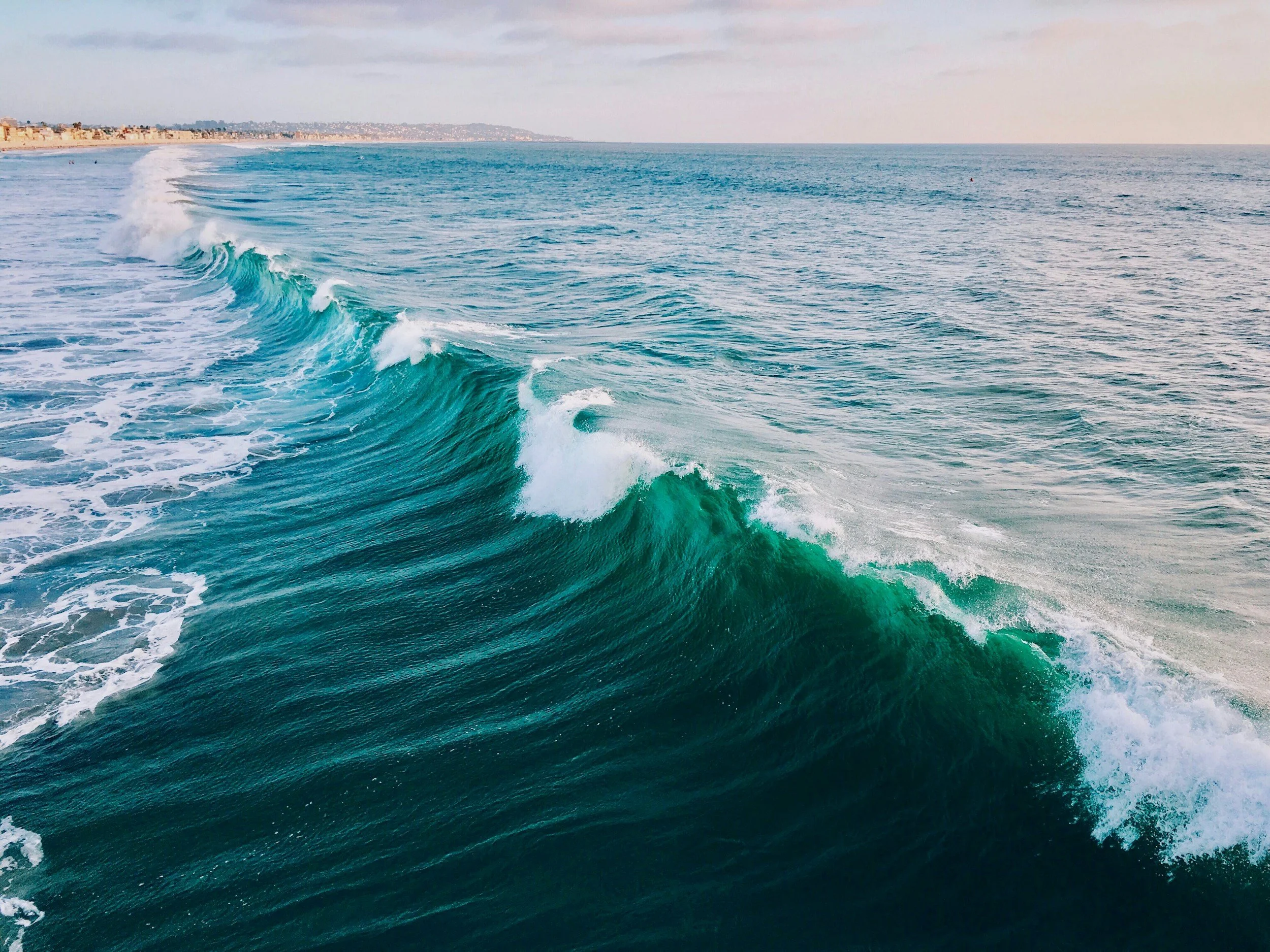 Ocean waves crashing near the shoreline with a distant beachfront and a cloudy sky.