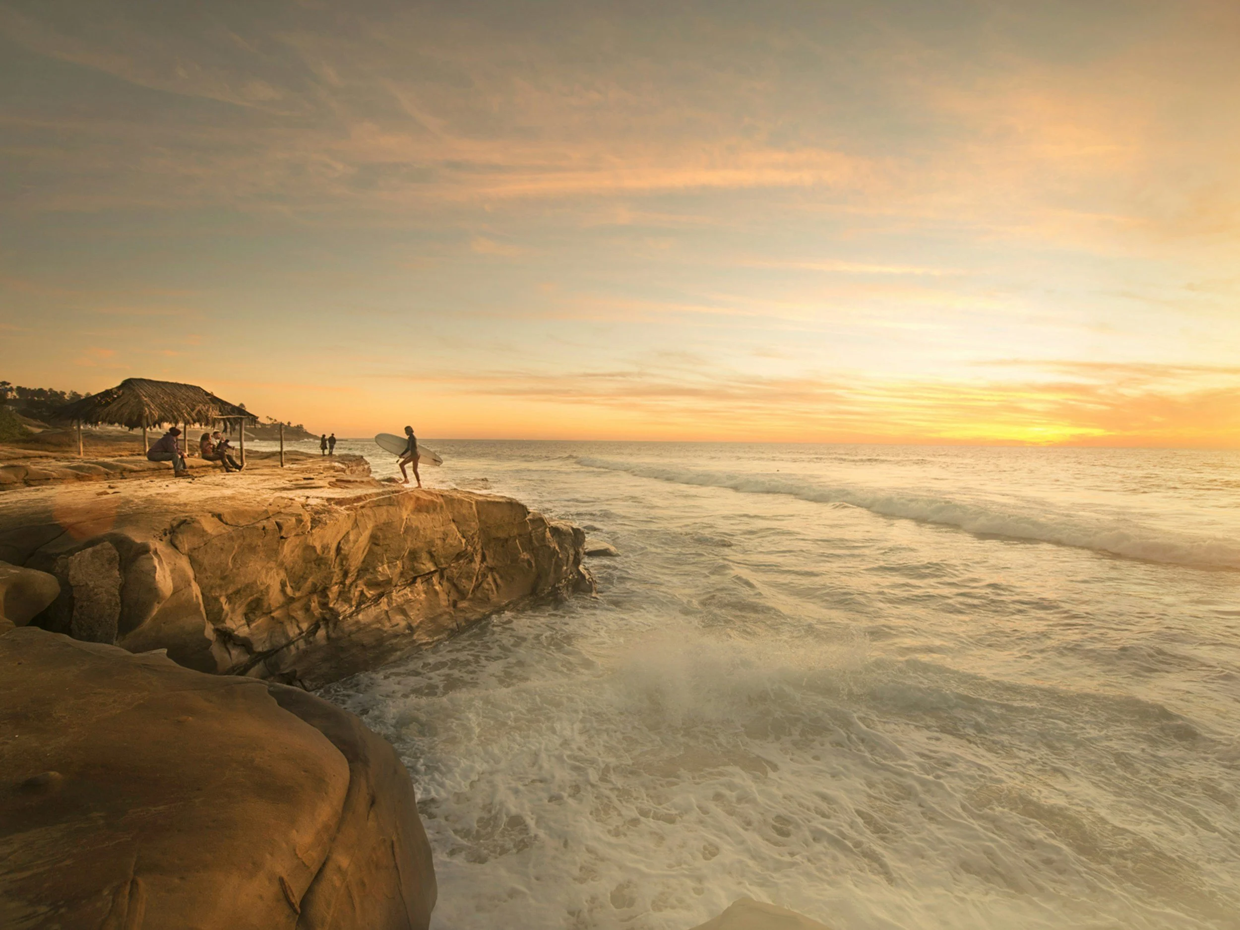 People on a rocky beach during sunset, some sitting under a thatched umbrella, one person carrying a surfboard, with the ocean waves and sky with orange and yellow hues.