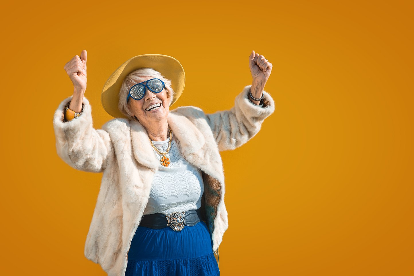 Senior woman in fur coat, gold chain, and jeweled sunglasses raising her arms in joy