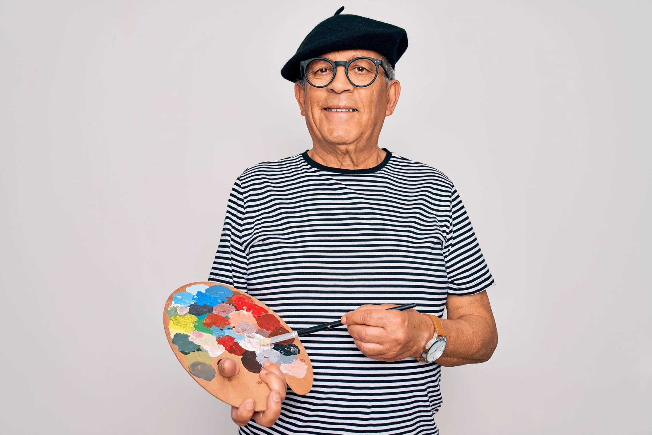 Older man wearing glasses, a black beret, and a striped shirt holding a painter's palette with various colors of paint and a brush in his right hand.