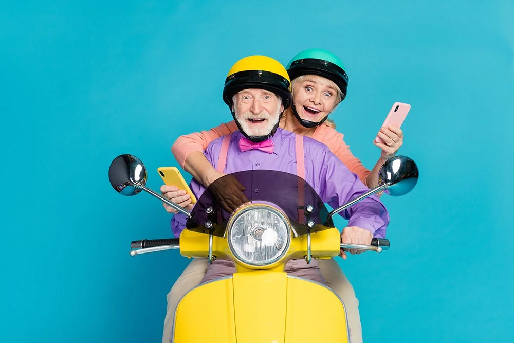 An elderly man and woman riding a yellow scooter, both wearing helmets and holding smartphones, with excited expressions against a blue background.