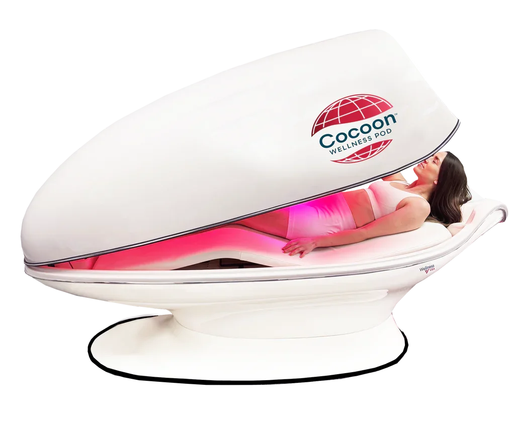 A woman lying inside a wellness pod with a curved lid that is partially open, displaying pink lighting inside. The wellness pod has the logo and name "Cocoon Wellness Pod" on its exterior.
