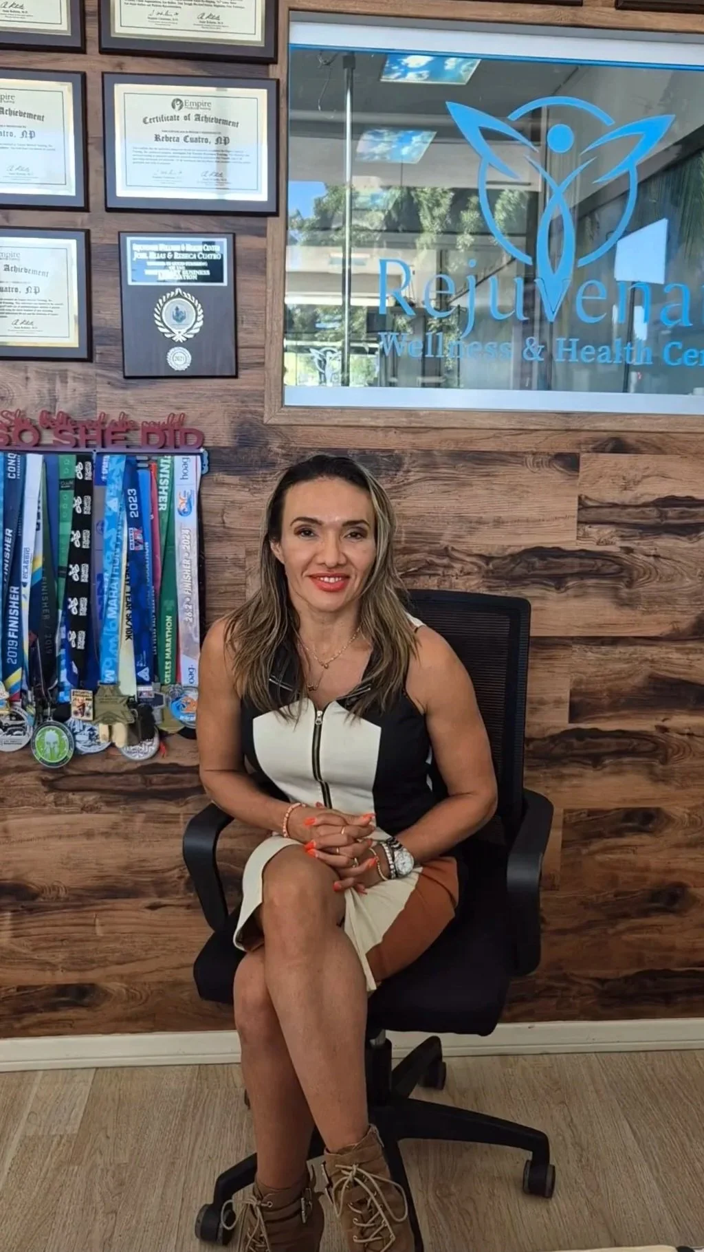 Woman sitting in an office chair at Rejuvena Wellness & Health Center, with medals hanging on a wall behind her and certificates on the wall.