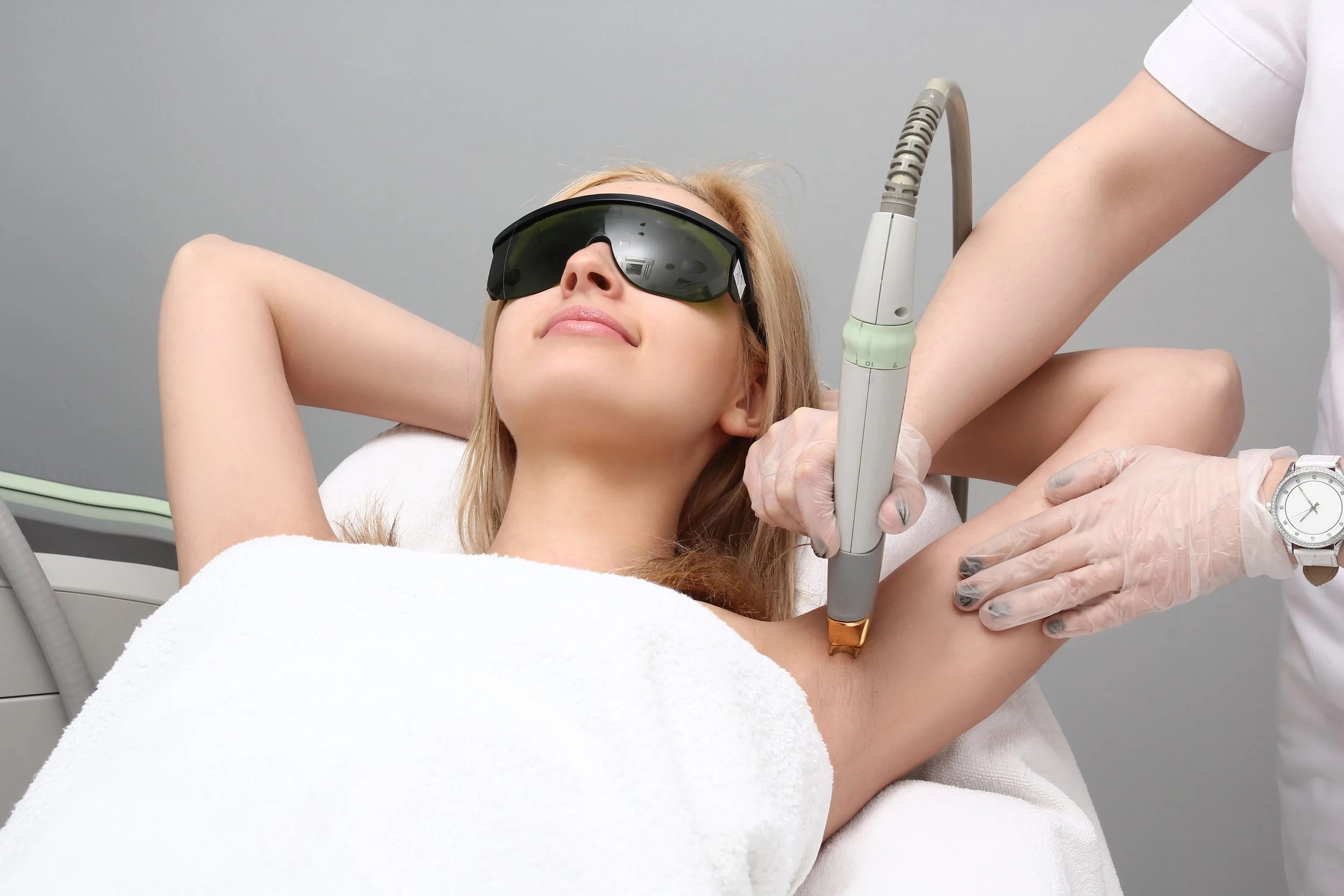 A woman is lying on a treatment bed, receiving laser hair removal on her armpit from a medical professional wearing gloves.