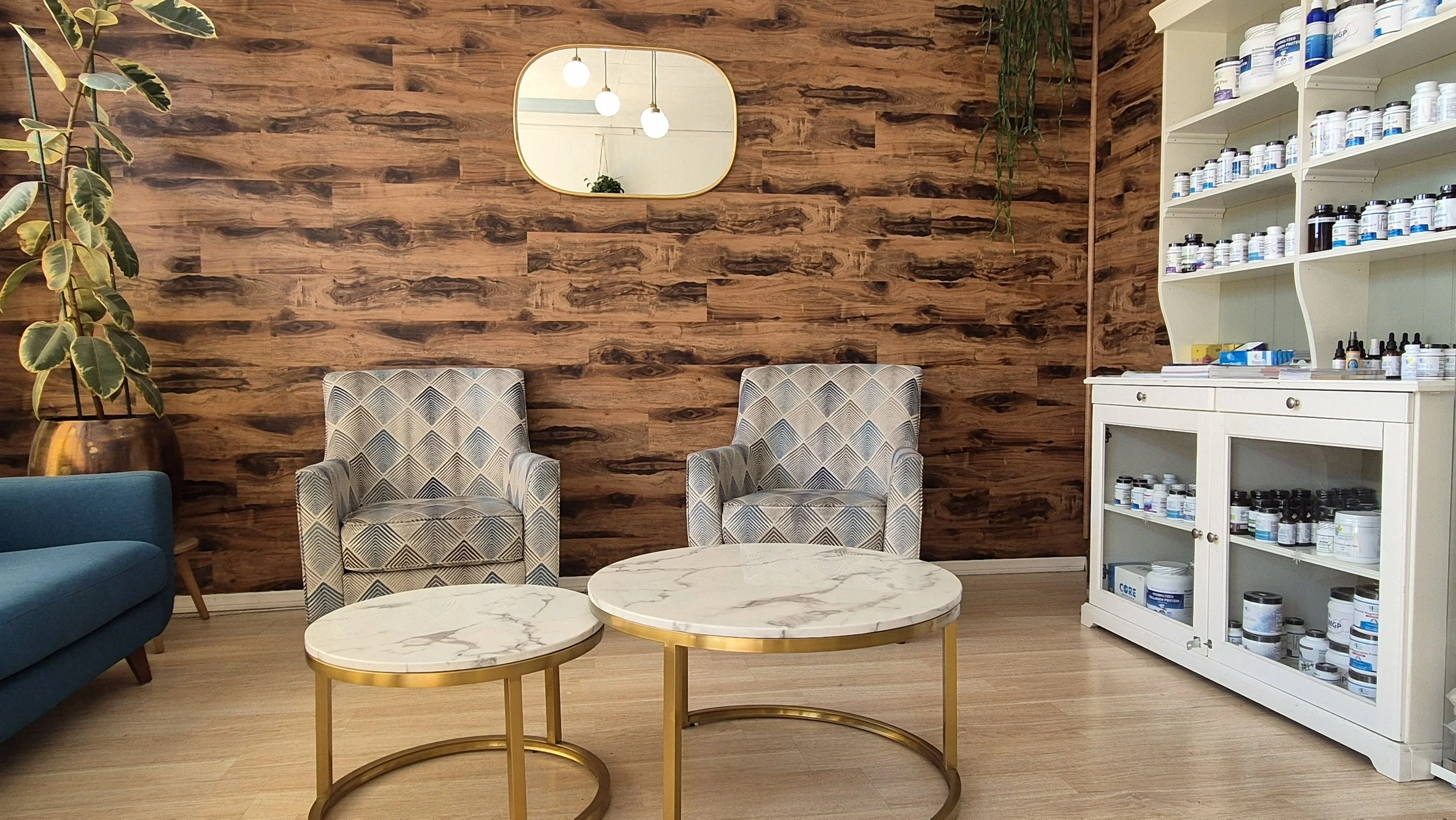 Waiting area with two patterned armchairs, a blue sofa, and two round marble-top coffee tables with gold bases. A large potted plant is on the left, a wooden wall with a mirror and hanging lights in the background, and a white cabinet with bottles of supplements or medication on the right.