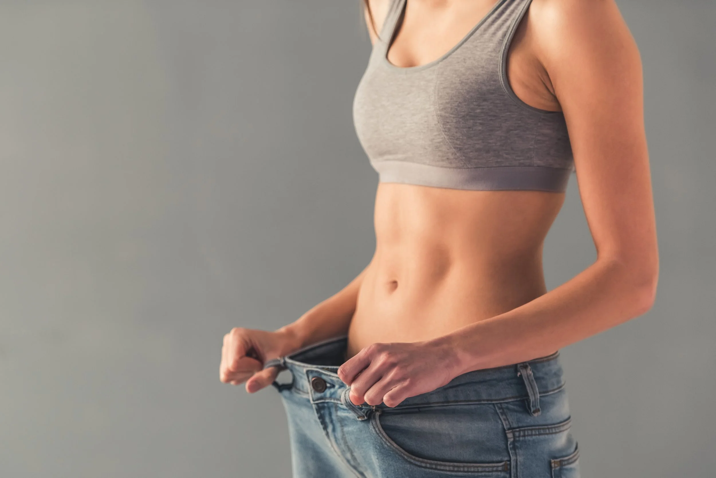 A woman holding loose fitting jeans, showing a fit physique with visible abdominal muscles, against a plain background.