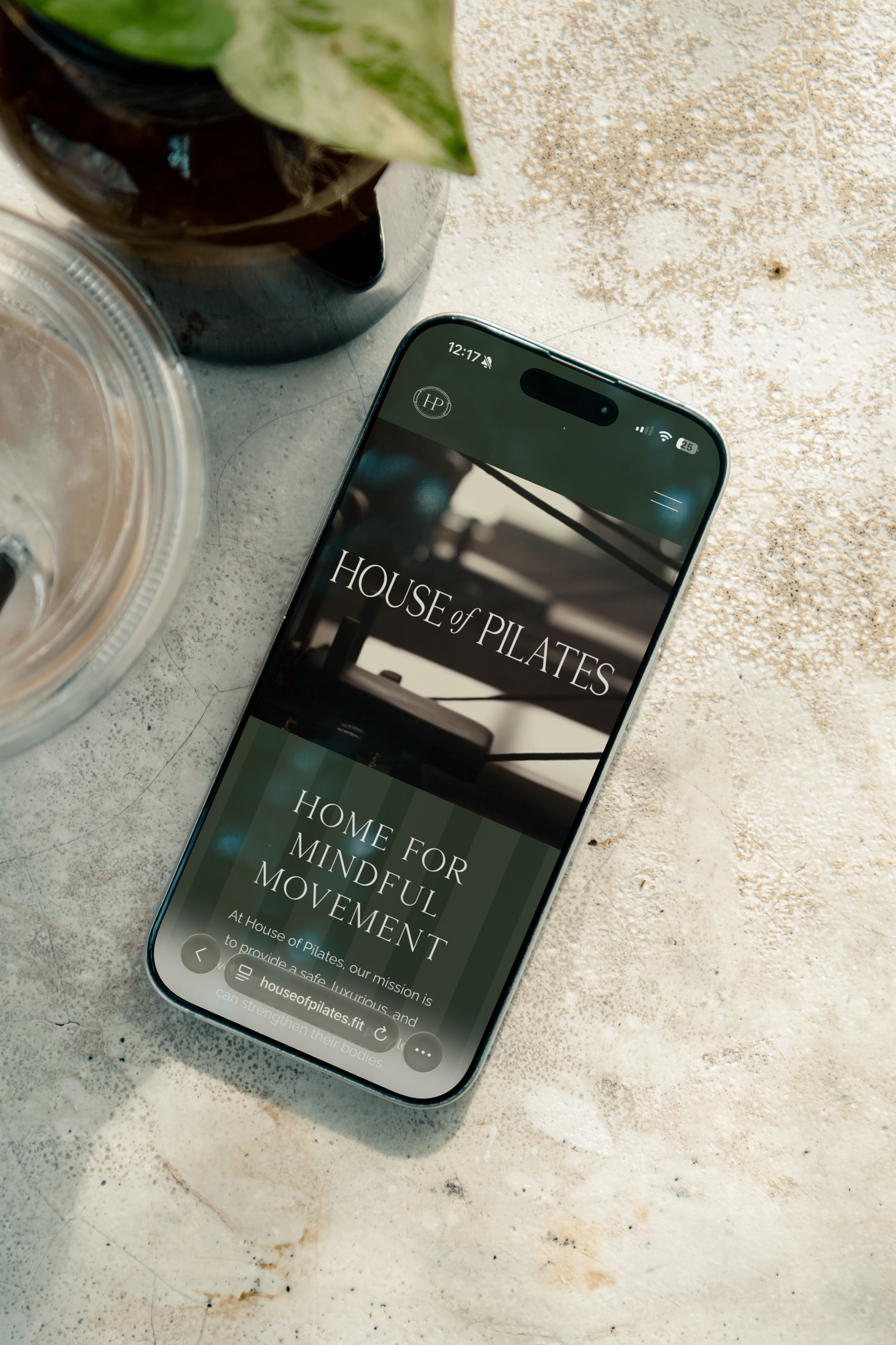 Smartphone displaying the homepage of 'House of Pilates' with a fitness theme on a light-colored textured surface, next to a plant and a glass jar with a lid.