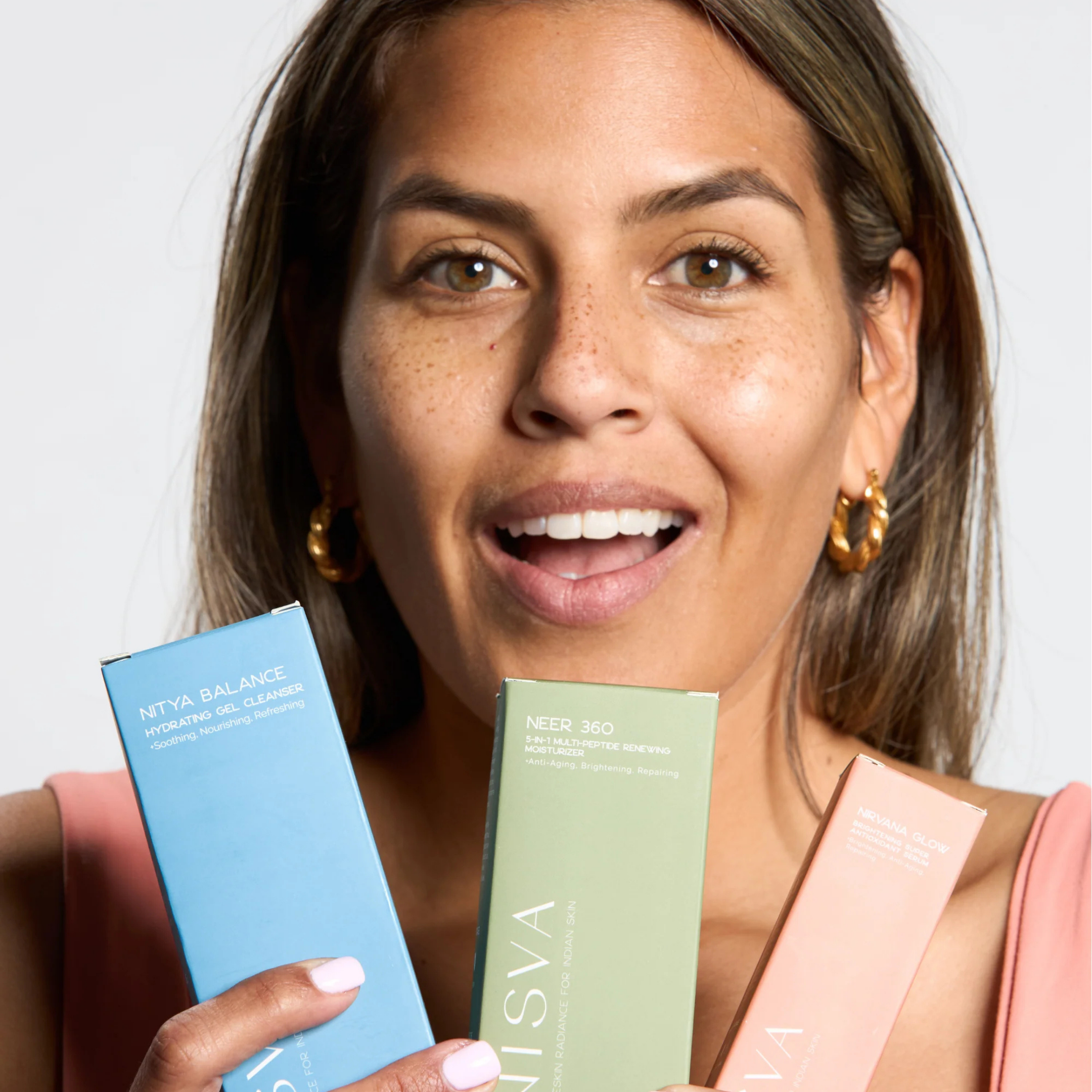 Woman holding three skincare product boxes, smiling, with gold hoop earrings and a pink top.