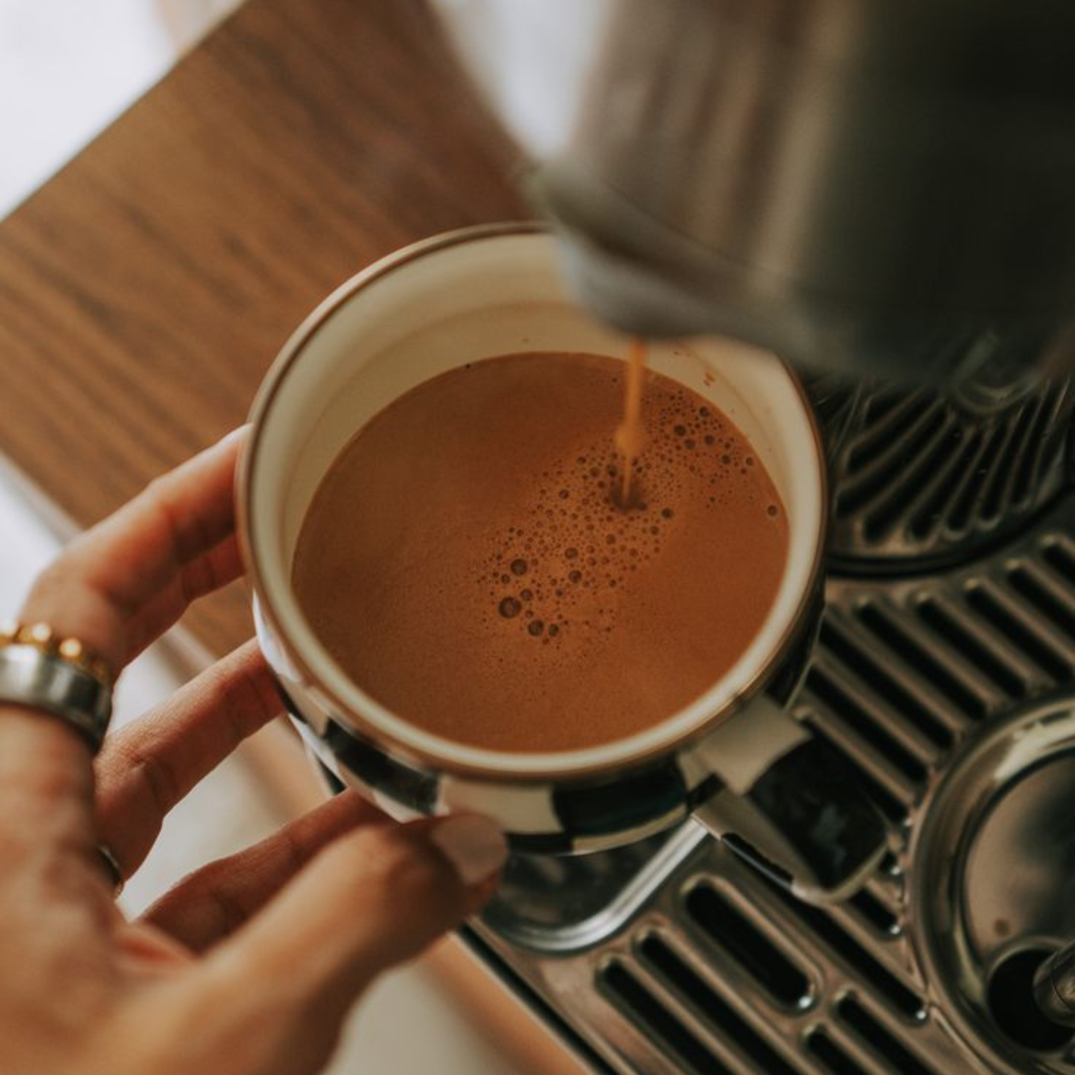 A person is pouring hot chocolate from a coffee machine into a mug, which is held by their left hand. The mug is ceramic with a dark exterior and a light interior. The person's hand has rings and a bracelet. The scene is on a wooden surface.