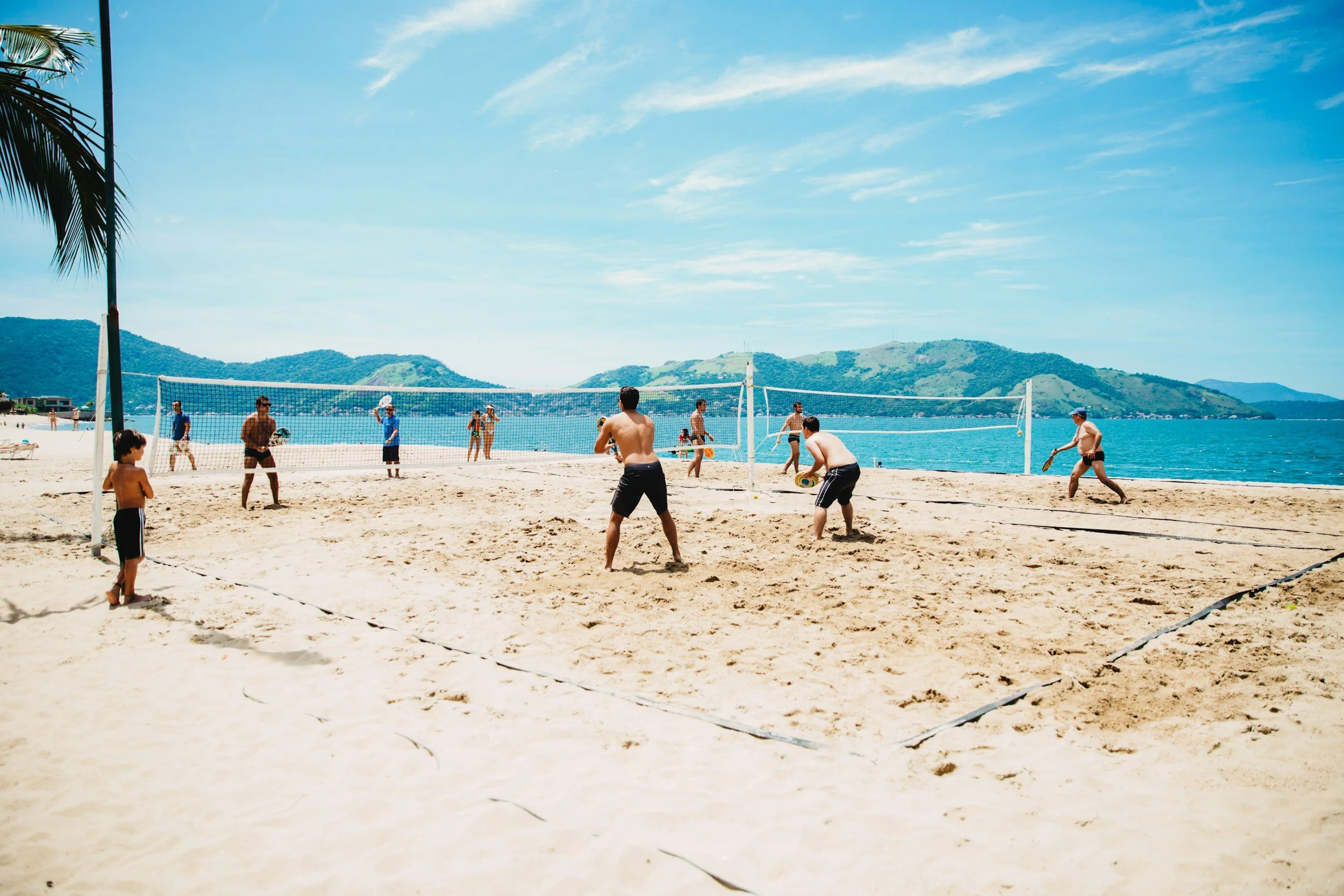 People playing beach volleyball on a sandy beach with mountains and water in the background.