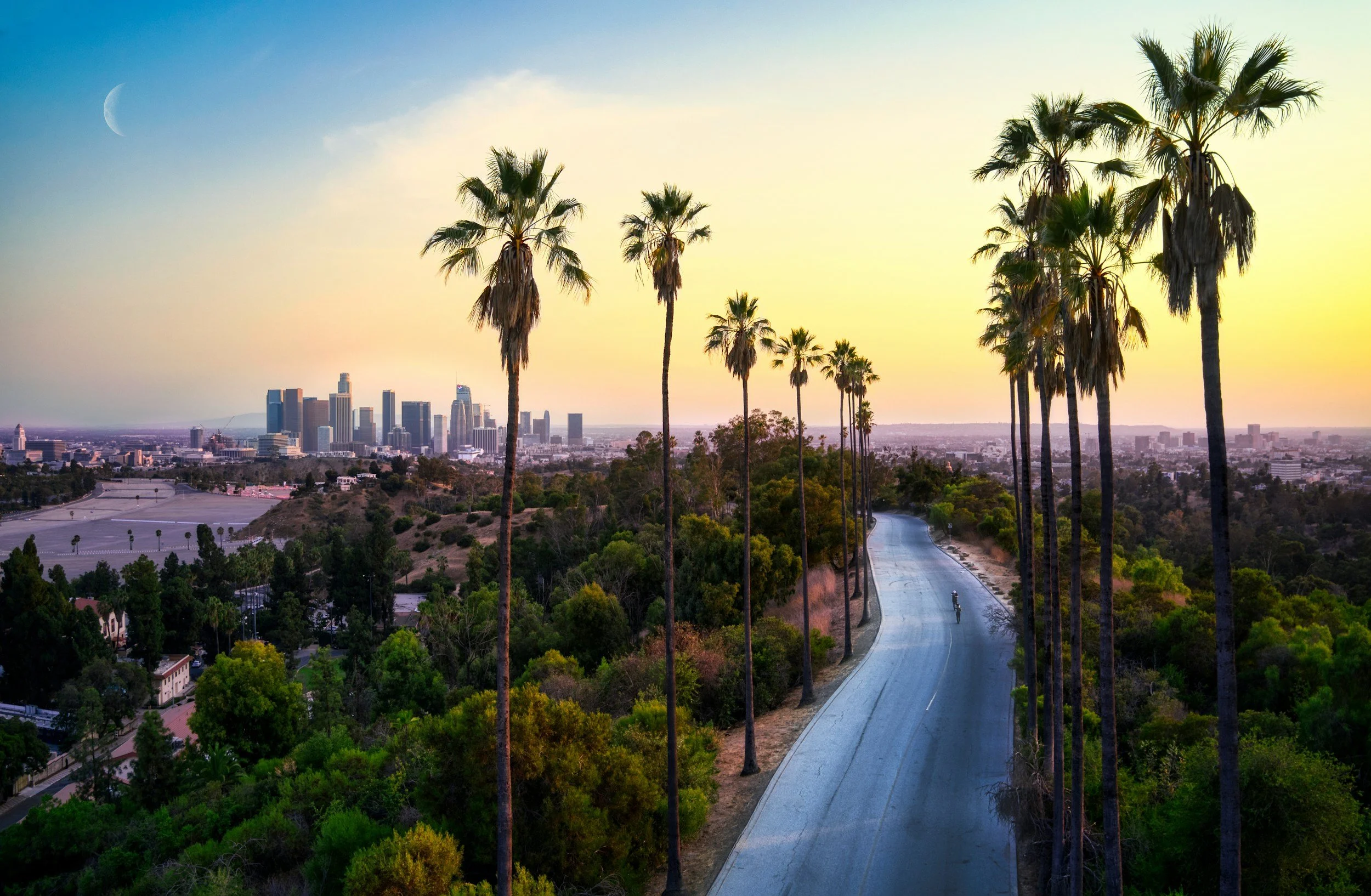 Sunset over a city skyline viewed from a winding road lined with tall palm trees, with the crescent moon visible in the sky.