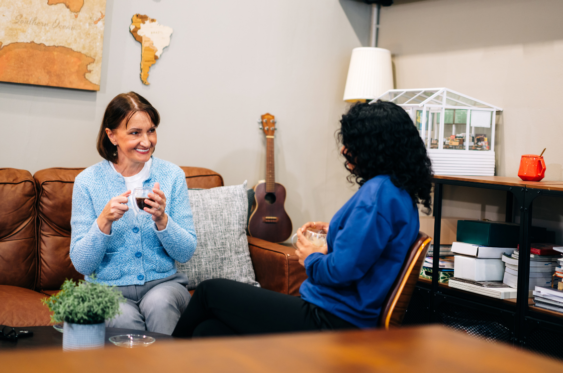 Two women having a conversation in a cozy living room, holding drinks, with a guitar and a world map on the wall behind them.