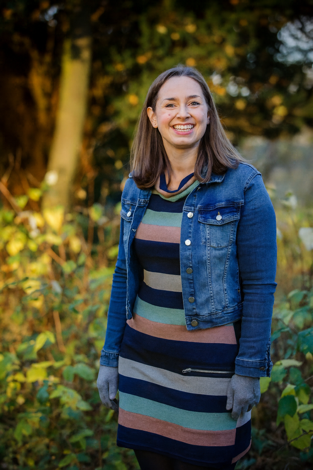 A woman with shoulder-length brown hair smiling outdoors during autumn, wearing a denim jacket over a striped multicolored dress, with gray gloves, surrounded by trees and foliage.