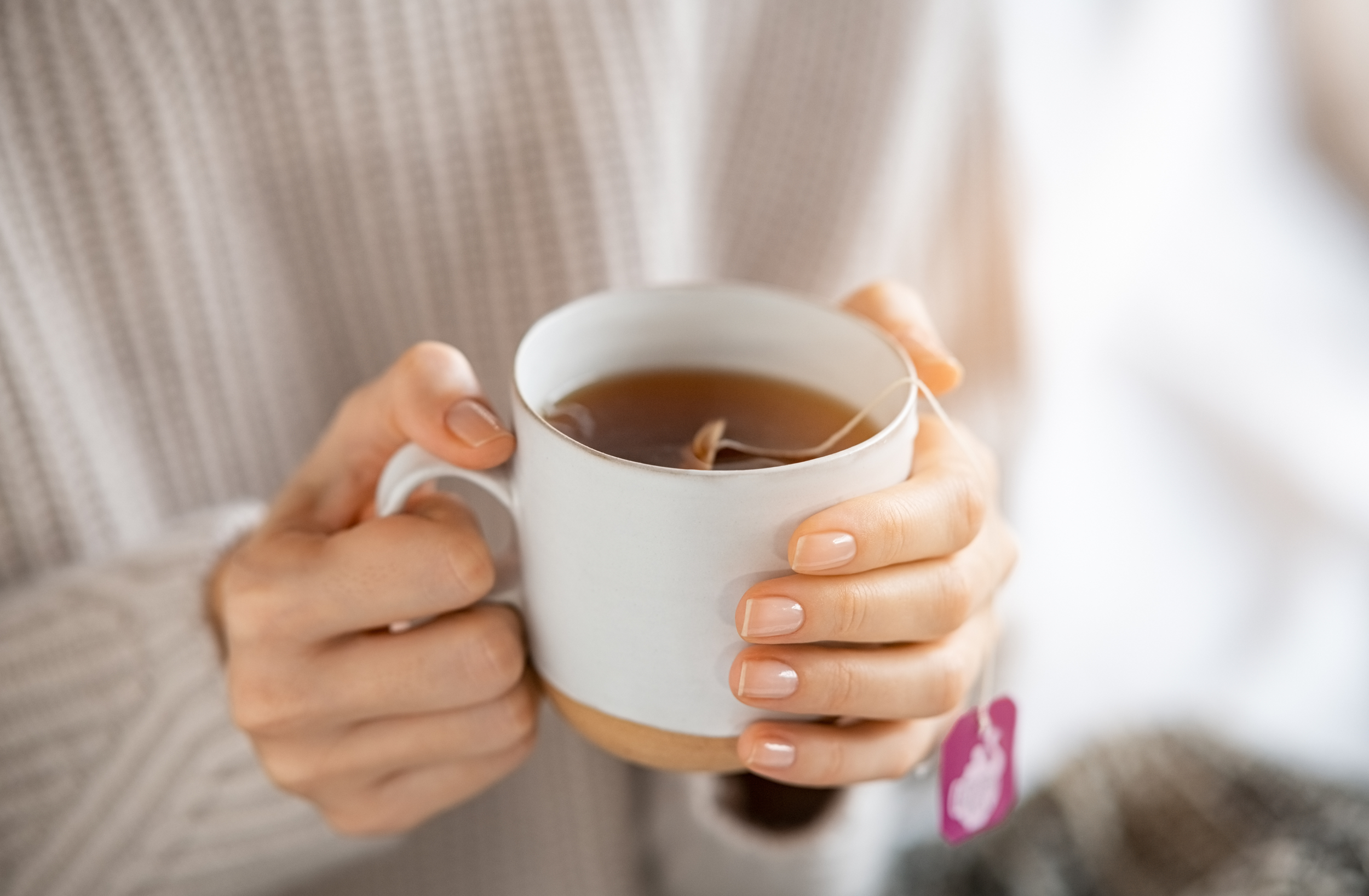 Person holding a white ceramic mug filled with tea, with a tea bag inside, in front of a beige knitted sweater.