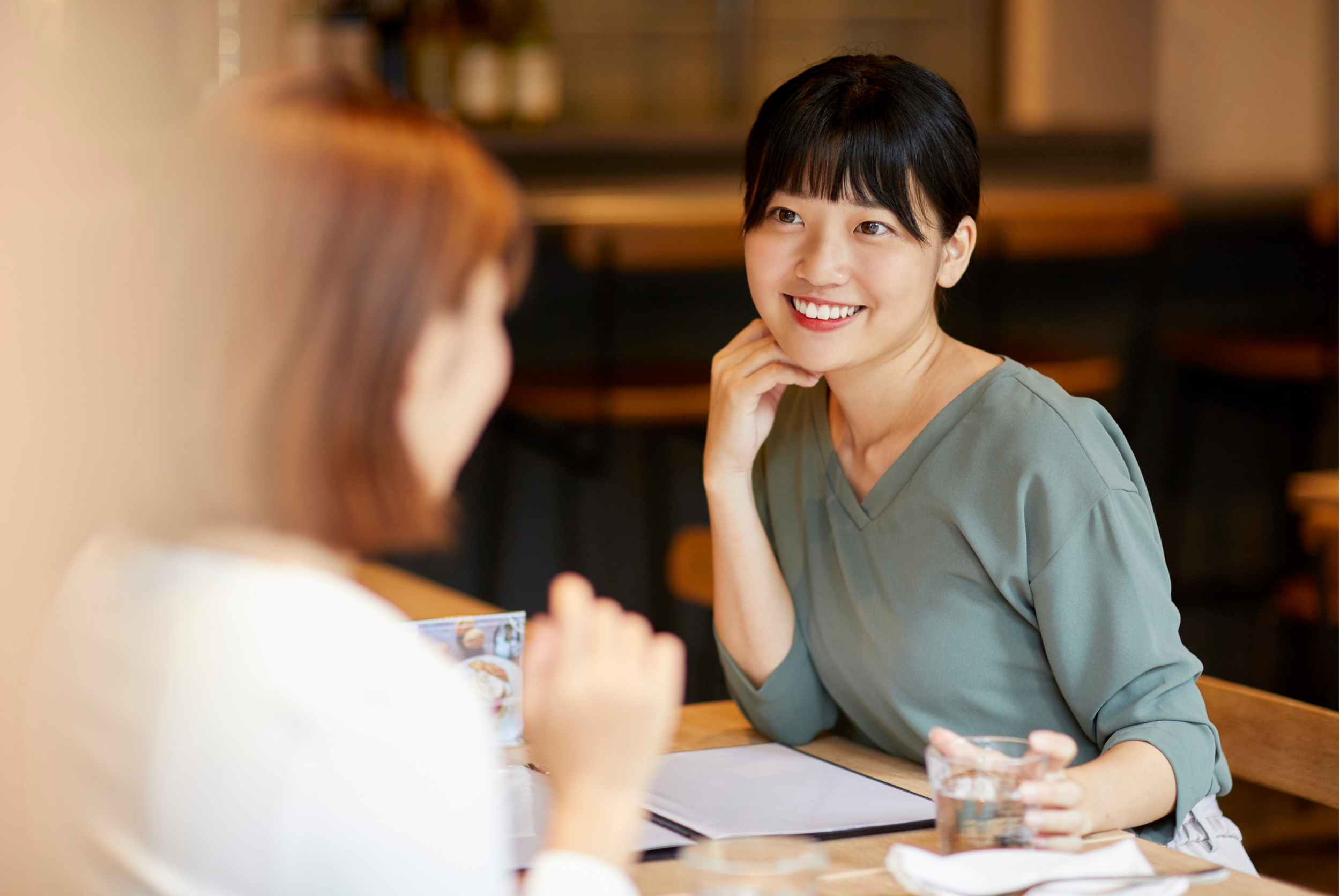 Two women sitting at a restaurant table, one smiling with her chin resting on her hand, the other slightly out of focus holding a menu or paper, with a folder and a glass of water on the table.