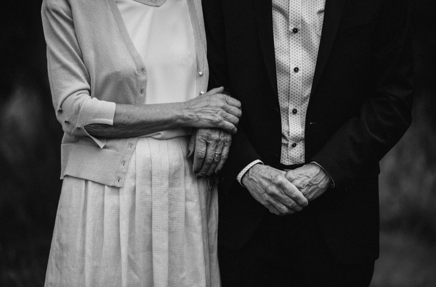 A black and white photo of an elderly woman and man holding hands, with the woman's hand resting on the man's arm.
