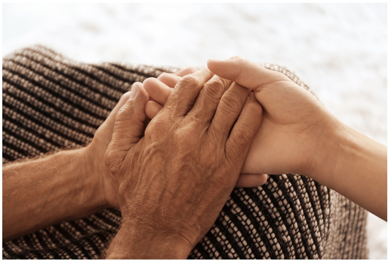 Two hands, one elderly and one young, clasped together gently, symbolizing care and compassion, with a textured brown and beige woven fabric in the background.