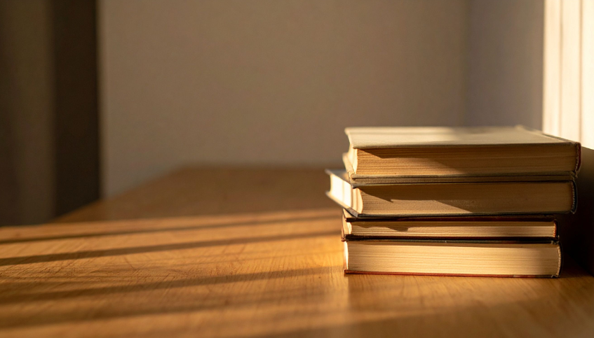 A stack of six books on a wooden surface with sunlight casting shadows.