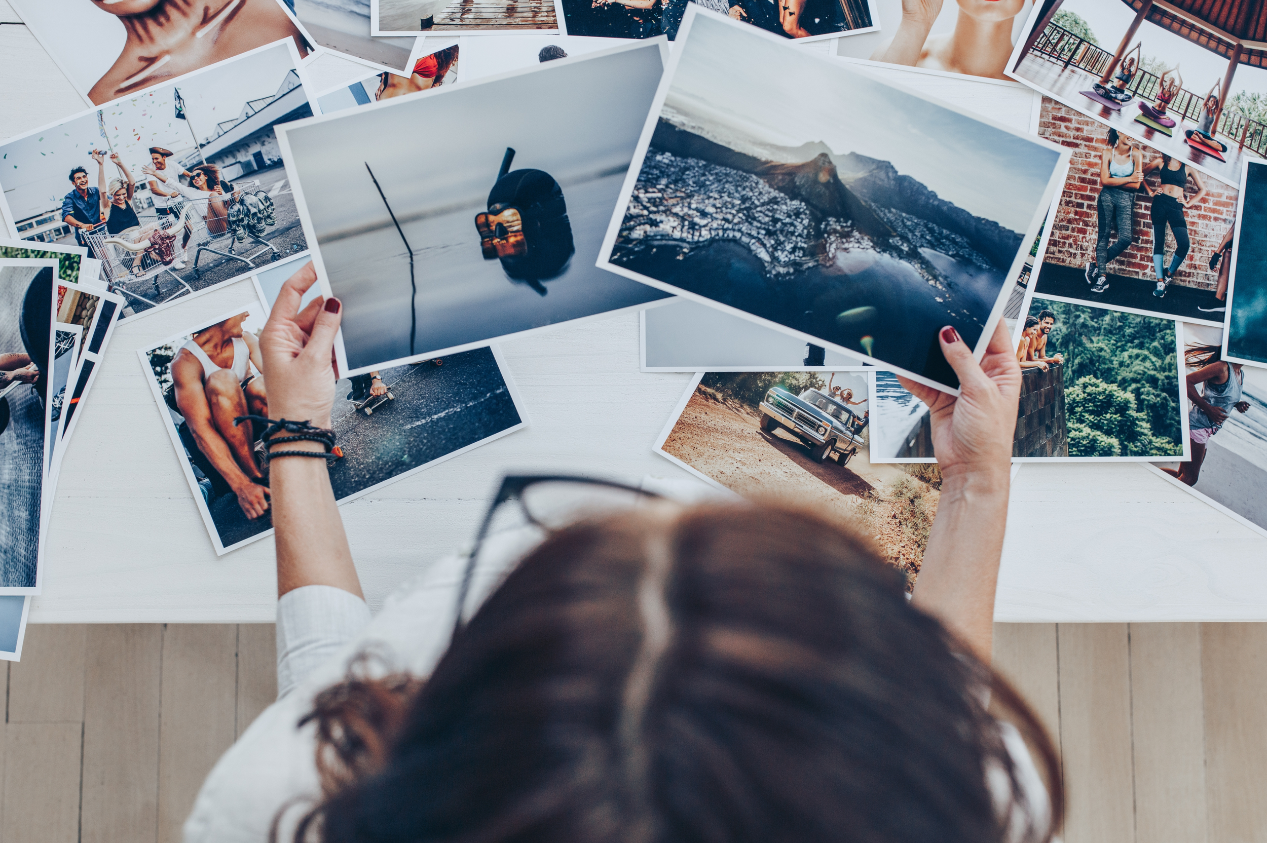 A person sitting at a white table surrounded by printed photographs, holding one with a landscape of mountains and a city, with various other photos scattered around.