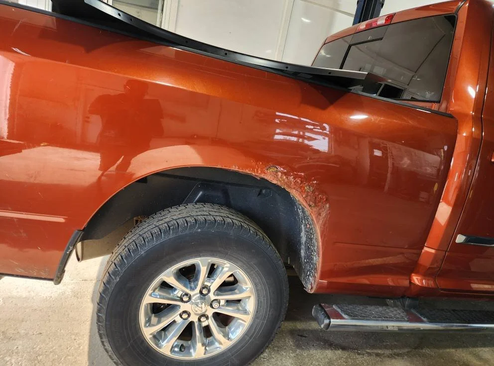 Close-up of the front passenger side of an orange pickup truck, showing damage with rust and scratches around the wheel well.