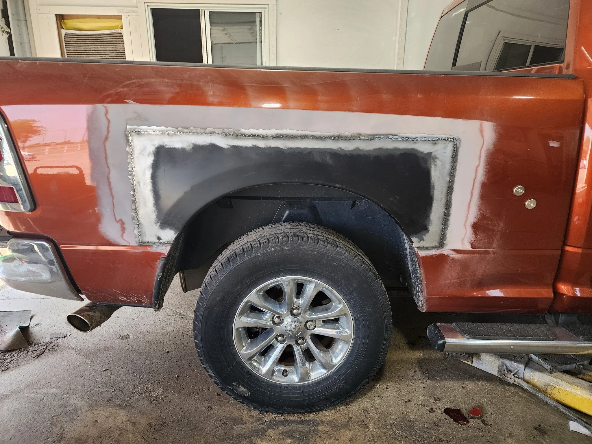 Side view of a pickup truck with a rust-colored body, showing a repair area on the rear quarter panel with primer and welding marks. The truck has a large tire and a step bumper, parked in a garage or workshop.