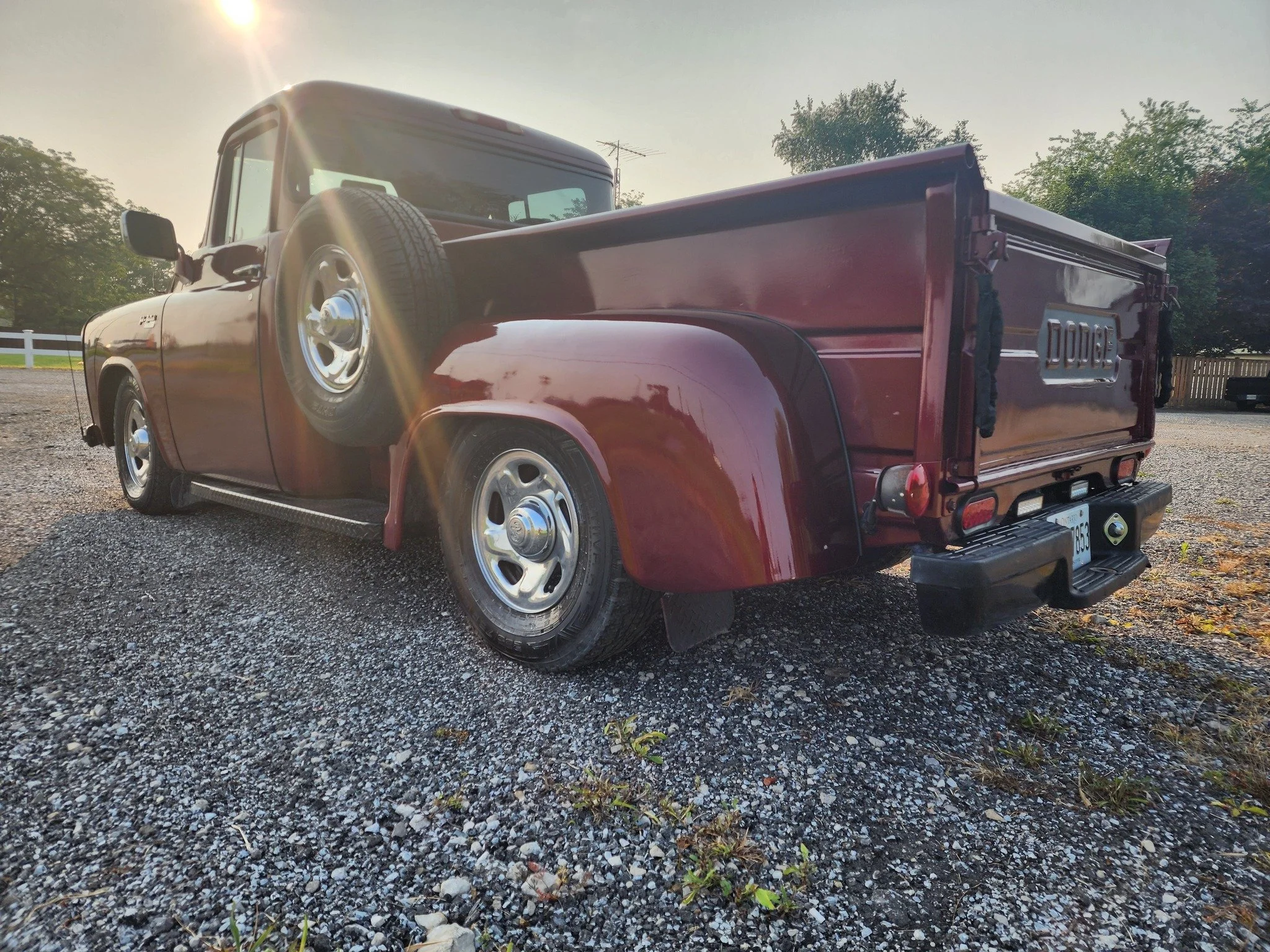 A vintage red Dodge pickup truck parked on gravel with the sun setting in the background.