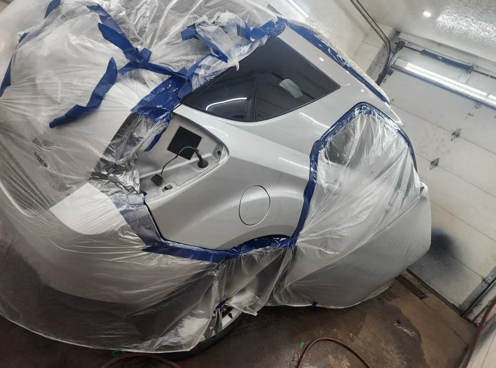 A white sports car inside a garage, covered with plastic sheeting and masking tape, likely in preparation for painting.